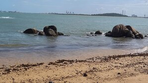 A beach with large rocks in the water, a sandy shore, and distant buildings.