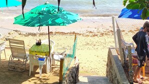 A beachside area with a table and chairs under a green umbrella, a stone wall, and a fence.