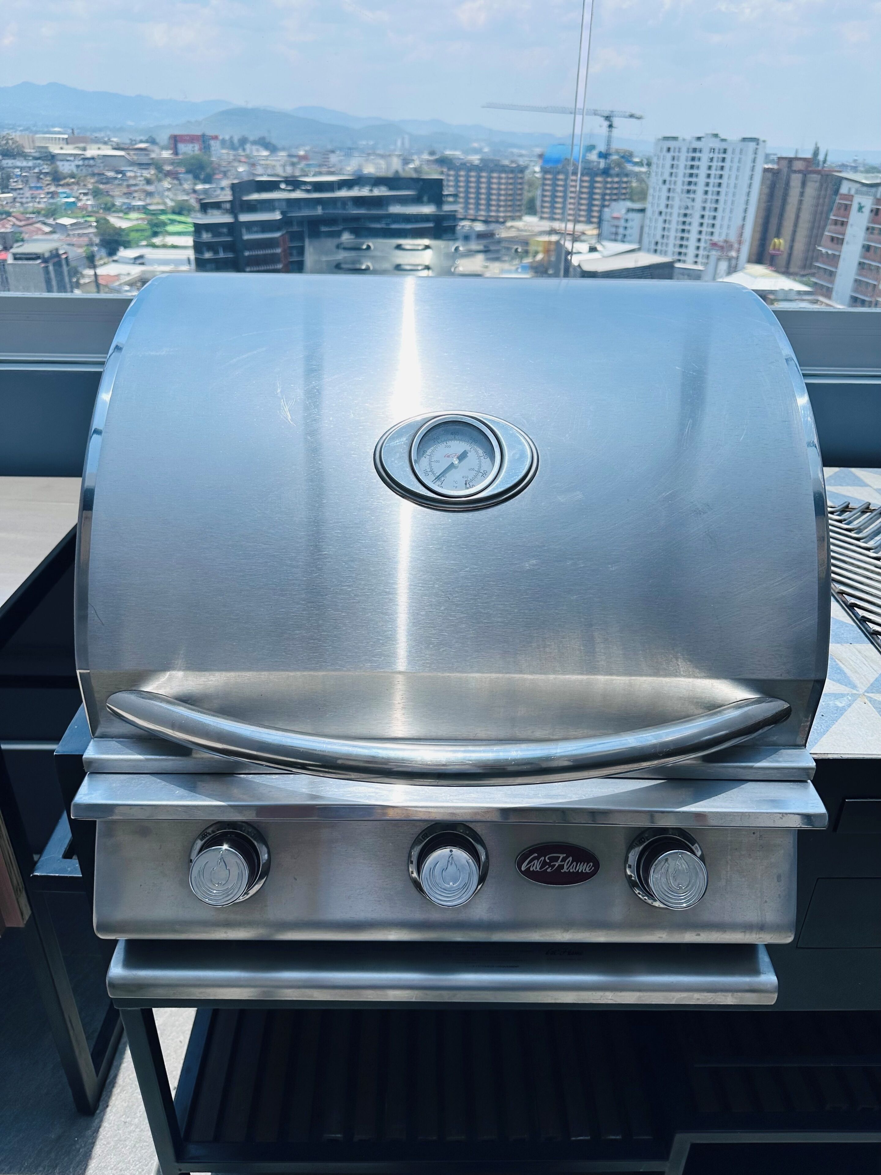 Una parrilla de acero inoxidable con un termómetro digital, situada en un balcón con vistas a la ciudad.