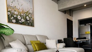 A modern living room with a sectional sofa, a round ottoman, a large painting, and a kitchen area visible through an open doorway. - Modern loft, excellent location and total comfort (Ciudad de Guatemala)