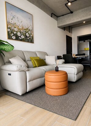 A modern living room with a sectional sofa, a round ottoman, a large painting, and a kitchen area visible through an open doorway. - Modern loft, excellent location and total comfort (Ciudad de Guatemala)