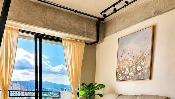 A modern living room with a leather sofa, a large window with curtains, a potted plant, and a city view. - Modern loft, excellent location and total comfort (Ciudad de Guatemala)