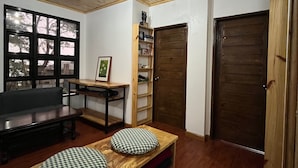 A room with a wooden ceiling, a large window, a wooden door, a sofa, a table with cushions, and a bookshelf.