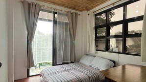 A bedroom with a wooden ceiling, a bed with striped bedding, a window with curtains, and a view of greenery outside.