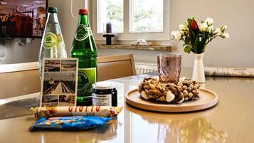 A dining table with a bottle of beer, a glass, a plate of nuts, a jar of jam, a rolled-up pastry, and a vase with flowers.
