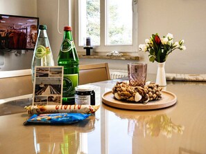 Ein Esstisch mit einer Bierflasche, einem Glas, einer Schale mit Nüssen, einem Marmeladenglas, einem gerolltem Gebäck und einer Vase mit Blumen.