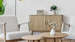 Ein modernes Wohnzimmer mit zwei runden Holztischen, einer weißen Vase mit Blumen, einem Holzschrank und einer großen Zimmerpflanze.