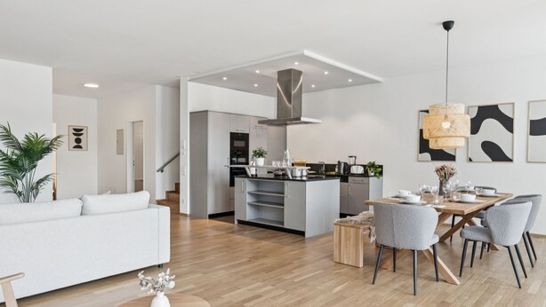 Ein modernes Wohnzimmer mit Sofa, Esstisch und Stühlen, eine Küche mit Kochinsel sowie eine Treppe zu einer weiteren Ebene.