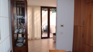 Una habitación con una mesa de comedor de madera, un aparador de vidrio con platos y vasos, y un ventanal corredero que da a un espacio exterior con una silla y una mesa.