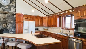 A kitchen with wooden cabinets, a stone wall, a large island with bar stools, a refrigerator, a sink, a dishwasher, and a clock on the wall.
