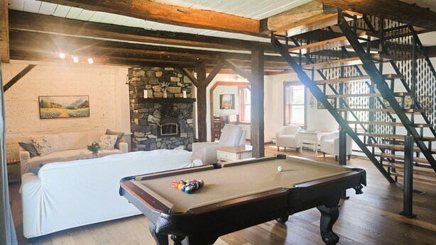 A pool table in a room with a stone fireplace, wooden beams, and a staircase.