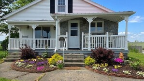 A two-story house with a porch, stone foundation, and a front yard with colorful flowers.
