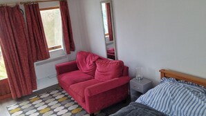 A room with a red sofa, a bed with striped bedding, a small bedside table with a lamp, a patterned rug, and a window with curtains.