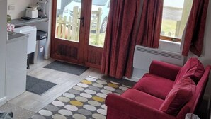 A compact room with a kitchenette, a red sofa, a patterned rug, and a view through a door.
