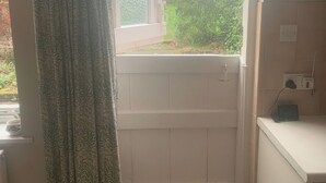 Une salle de bain avec une douche, une fenêtre avec des rideaux, un radiateur et une petite étagère sur laquelle se trouve un appareil.