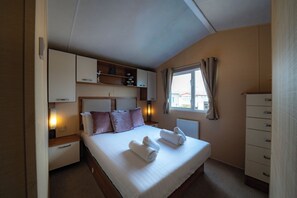 Ein Schlafzimmer mit Bett, Kissen, einem Fenster mit Vorhängen, einem Schminktisch und einem Regal mit Gegenständen.