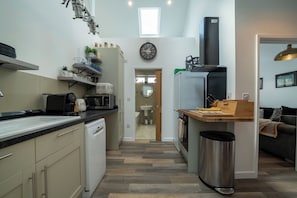 Eine kompakte Küche mit Geschirrspüler, Mikrowelle und kleinem Kühlschrank. Ein Holztisch mit Messerblock und Mülleimer steht dort. An der Wand sind eine Uhr und ein Fenster zu sehen.