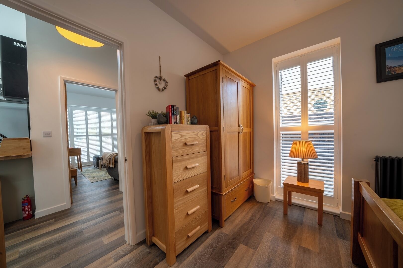 Une pièce avec un meuble en bois, une petite table avec une lampe et une fenêtre avec des stores.