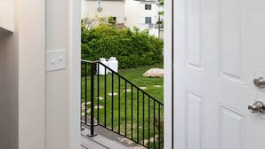 A white door with a black doormat, a small porch with a black railing, and a view of a green lawn and a house in the background. - Comfortable 3BR/2BA Near Old Greenwich & Stamford (Stamford)