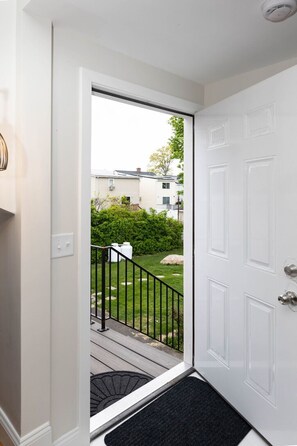 A white door with a black doormat, a small porch with a black railing, and a view of a green lawn and a house in the background.