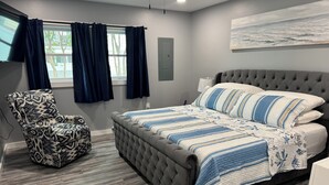 A bedroom with a tufted headboard bed, a patterned armchair, and a view of the outdoors through a window with curtains. - Downtown Rehoboth Ave Renovated Condo Pet Friendly (Rehoboth Beach)
