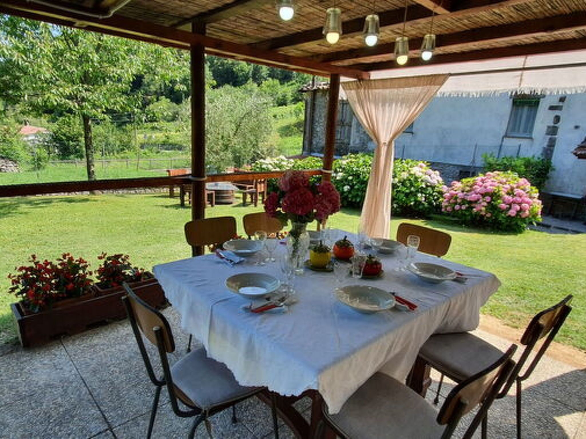 Un espace de restauration en plein air avec une table dressée pour quatre personnes, entourée de chaises, sous un toit abrité d’où l’on peut admirer un jardin et un bâtiment.