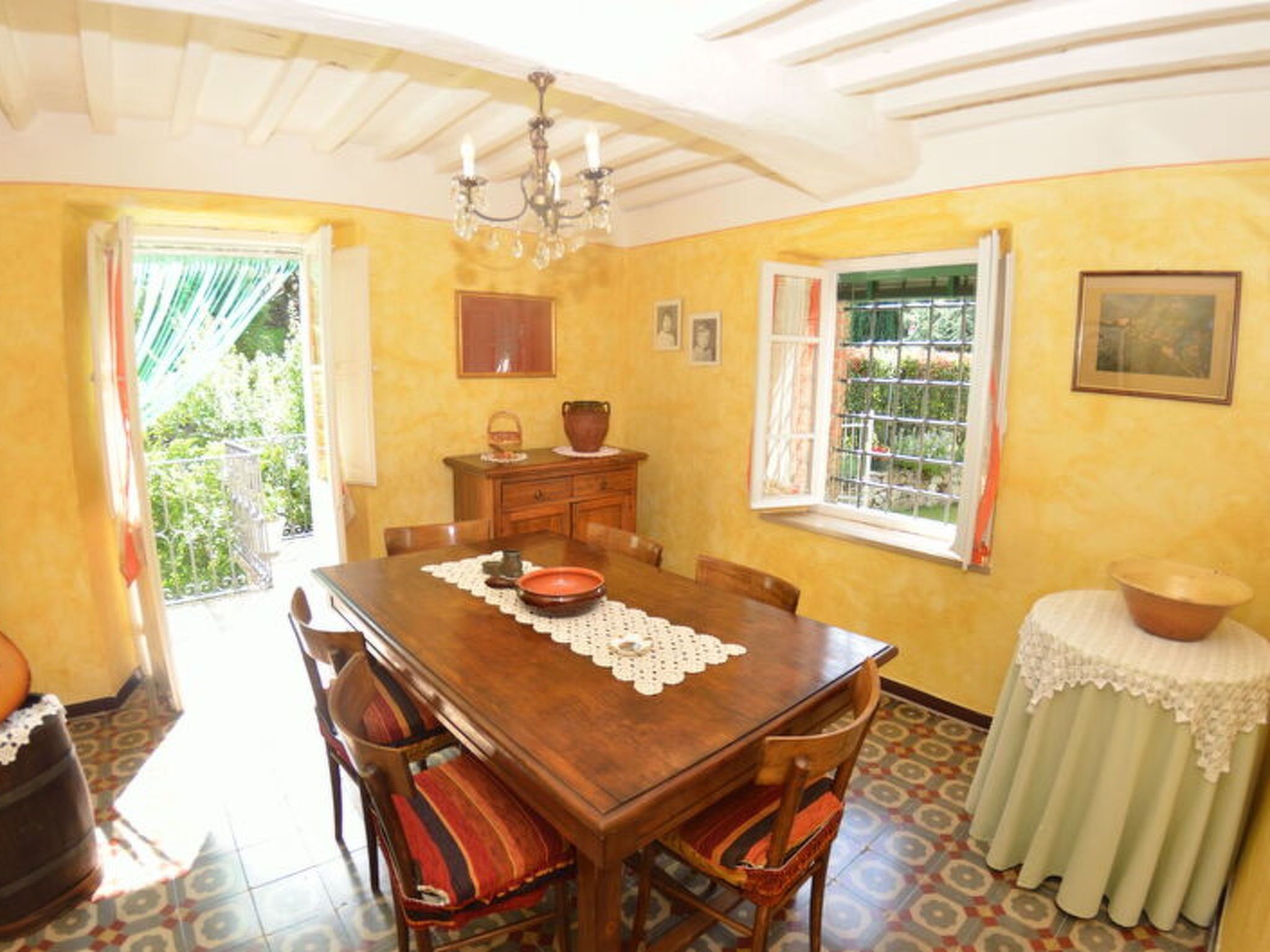A dining room with a wooden table, chairs, a chandelier, a window with a view, and a small table with a bowl.