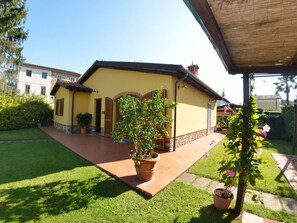 Une maison jaune avec un toit en tuiles, un jardin avec une terrasse dallée et un pot de fleurs au premier plan.
