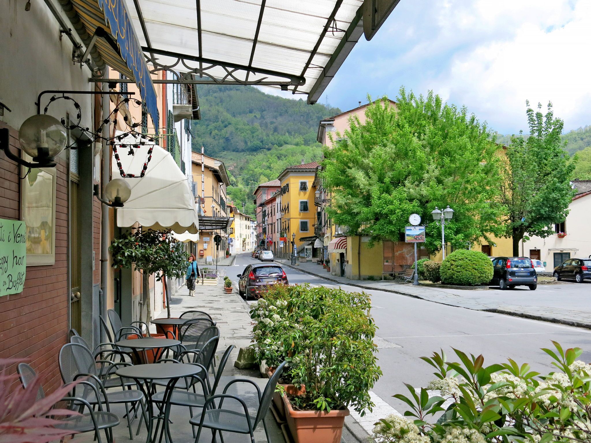 Une vue d’une rue avec des terrasses, une chaîne de montagnes en arrière-plan et des voitures garées.