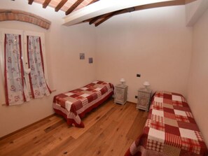 Ein Schlafzimmer mit zwei Einzelbetten, Holzboden, einem Fenster mit Vorhängen und einem kleinen Nachttisch mit Lampe.