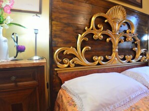 Ein hölzerner Kopfseitenverzierer mit aufwendigen Schnitzereien, ein Nachttisch mit Blumenvase und ein Bett mit floralem Überwurf.