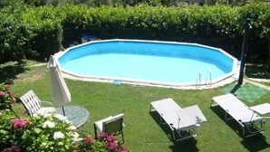 Ein rechteckiger Pool mit weißem Sonnendeck, umgeben von einem gepflegten Rasen und einer Hecke. Liegestühle und ein Tisch mit Glasplatte sind vorhanden.