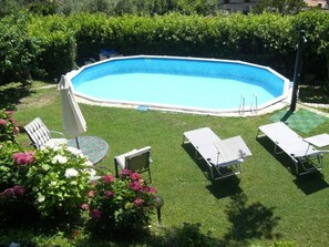 Ein rechteckiger Pool mit weißem Sonnendeck, umgeben von einem gepflegten Rasen und einer Hecke. Liegestühle und ein Tisch mit Glasplatte sind vorhanden.