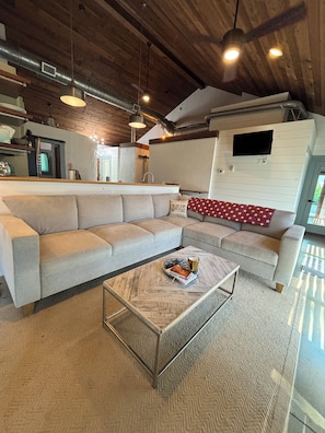 A modern living room with a sectional sofa, a coffee table, pendant lighting, and a visible kitchen area.