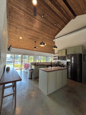 A modern kitchen with wooden ceiling beams, a central island, and large windows overlooking a water body.