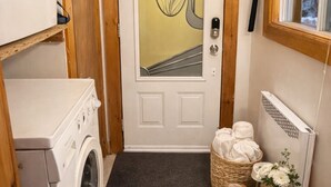 Uma lavanderia compacta com máquina de lavar, secadora, porta com painel de vidro decorativo, radiador e janela com vista para árvores.