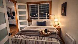 Quarto com cama, mesa de cabeceira, abajur e janela com persianas.