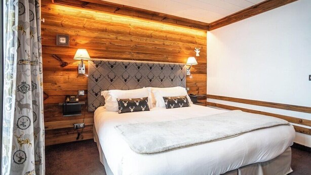 A bedroom with a wooden headboard, two bedside lamps, a bed with white bedding, and a nightstand with a lamp. - Les Campanules Hotel Restaurant (Tignes)