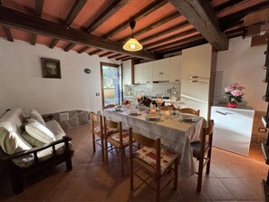 Una zona de menjador amb taula ben posada per a quatre, cadires de fusta, un sofà i una cuina al fons.