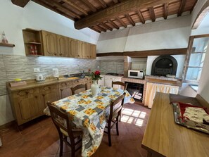 Une cuisine avec des meubles en bois, un dosseret carrelé, une table recouverte d’une nappe à motifs floraux et un poêle à bois.