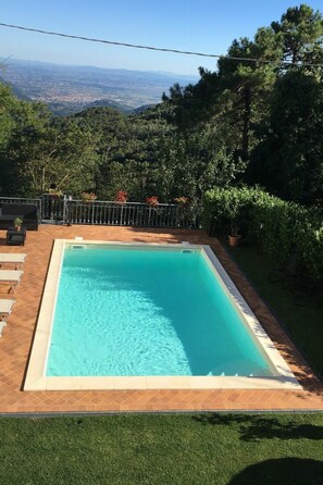 A rectangular swimming pool with a clear blue water, surrounded by a tiled deck, a grassy area in the foreground, and a view of a distant cityscape through the trees. - Borgo Di Zacco by Interhome (La Sassa)