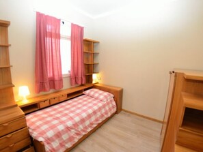 Una habitación pequeña con cama individual, estructura de madera, mesita de noche con lámpara, ventana con cortinas rosadas y estantería empotrada.