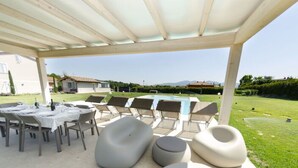 Un patio exterior con zona de estar cubierta, piscina y vistas al paisaje circundante.