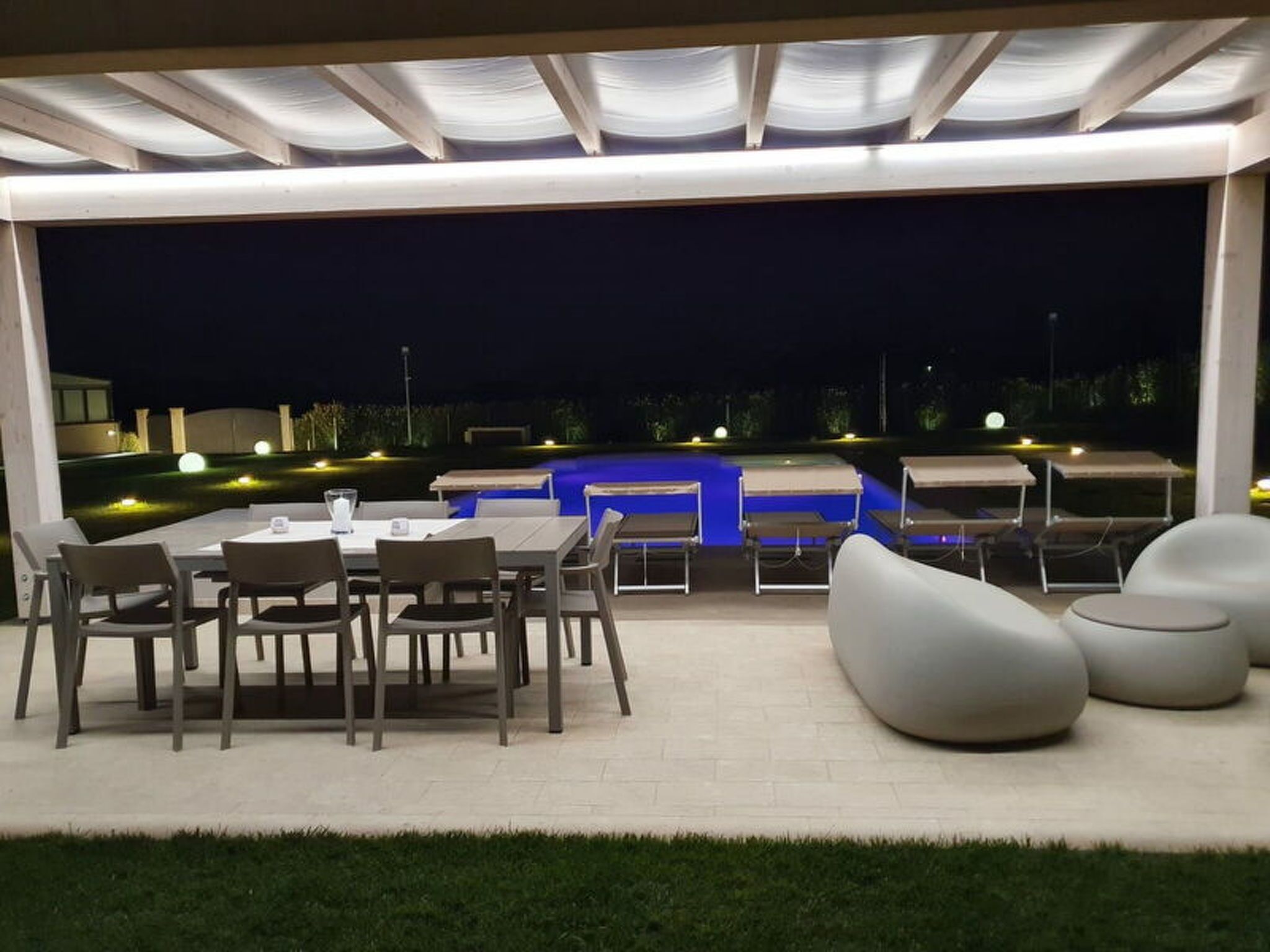 Uma área de pátio externa com mesas e cadeiras, uma piscina iluminada em azul e vista para a paisagem ao redor.