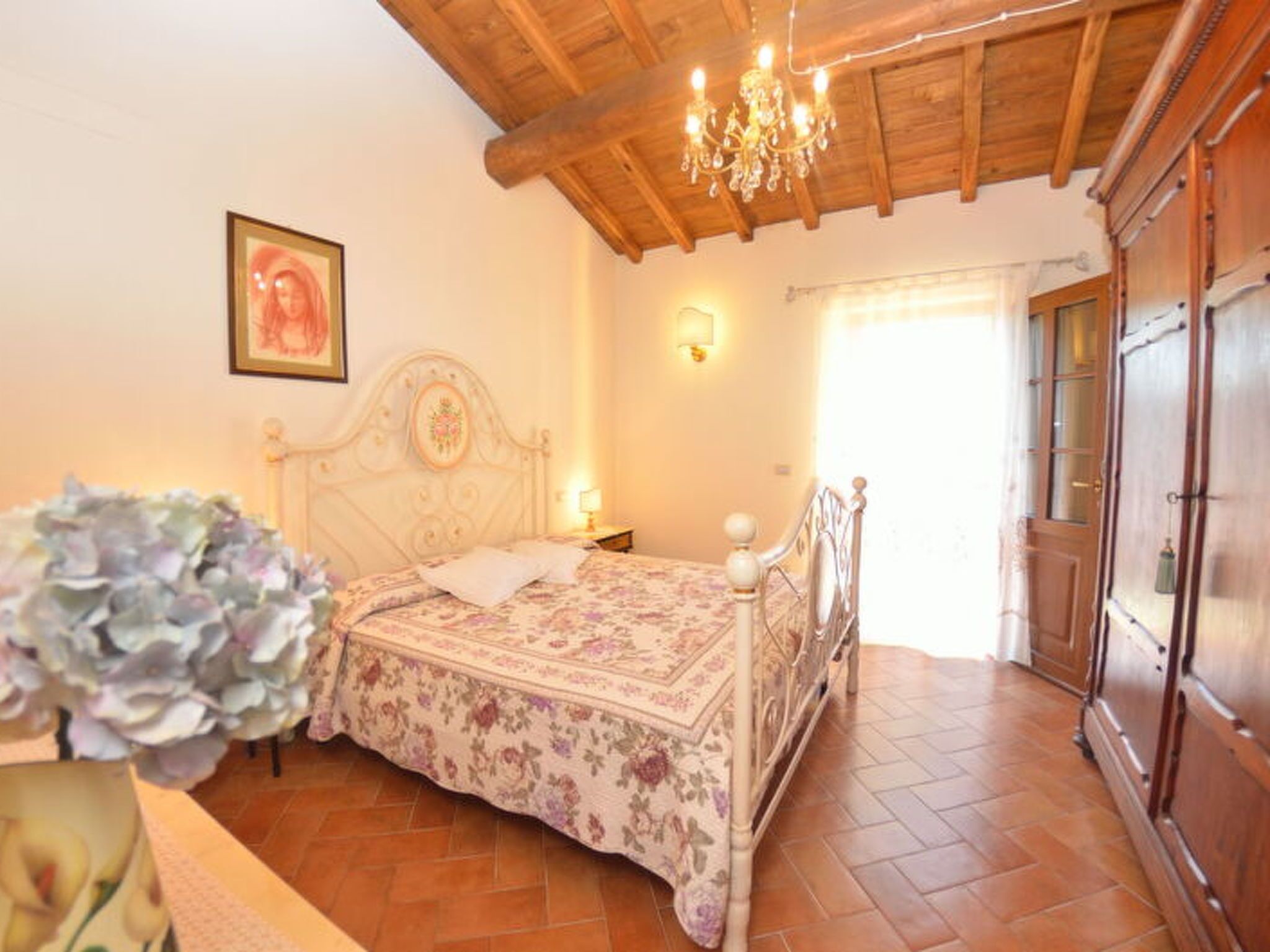 A bedroom with a wooden ceiling, a bed with a floral bedspread, a chandelier, a wooden wardrobe, and a window with curtains.