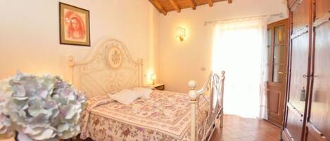 A bedroom with a wooden ceiling, a bed with a floral bedspread, a chandelier, a wooden wardrobe, and a window with curtains.