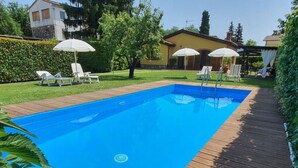 Una piscina rettangolare con un pontile in legno tutt'intorno, una zona verdeggiante con alberi e un edificio sullo sfondo.