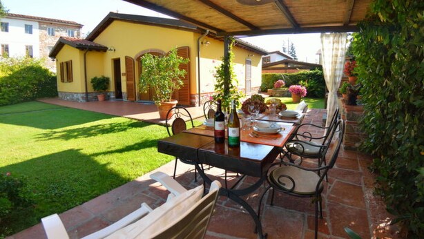 Patio esterno con tavolo apparecchiato per mangiare, circondato da sedie e una zona coperta con una ventola a soffitto.
