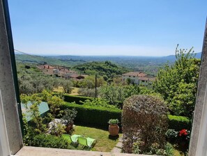 Blick aus einem Fenster auf einen üppigen Garten mit einem Weg, einer kleinen Sitzgelegenheit und entfernte Gebäude.