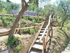 Eine Holztreppe mit Geländern führt einen Hang hinauf, umgeben von Grünpflanzen und einem Zaun.